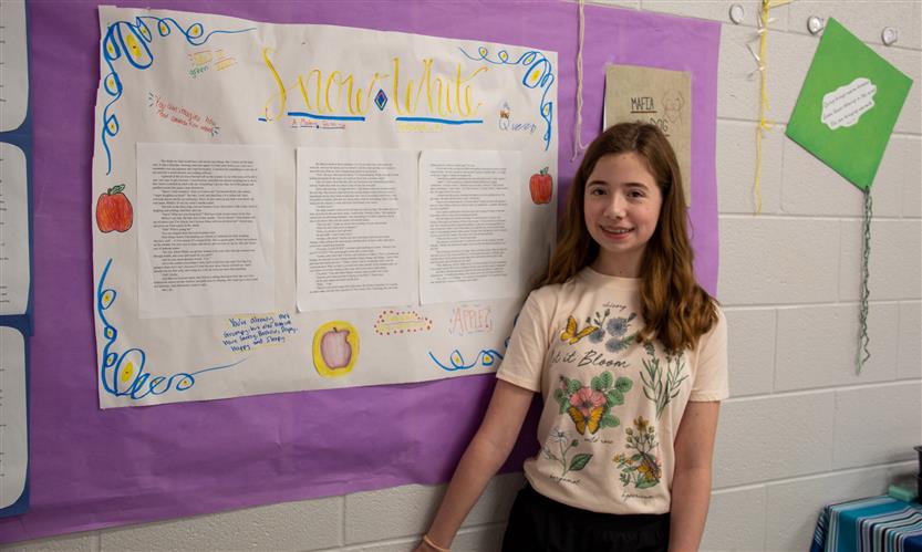 A girl with long, brown hair smiles while standing against a wall in a hallway